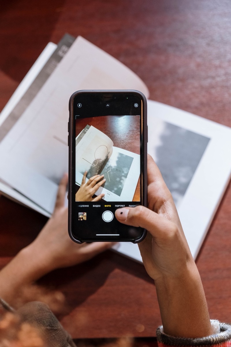 Руки держат смартфон над раскрытой книгой и фотографируют страницу, на экране видно приложение камеры